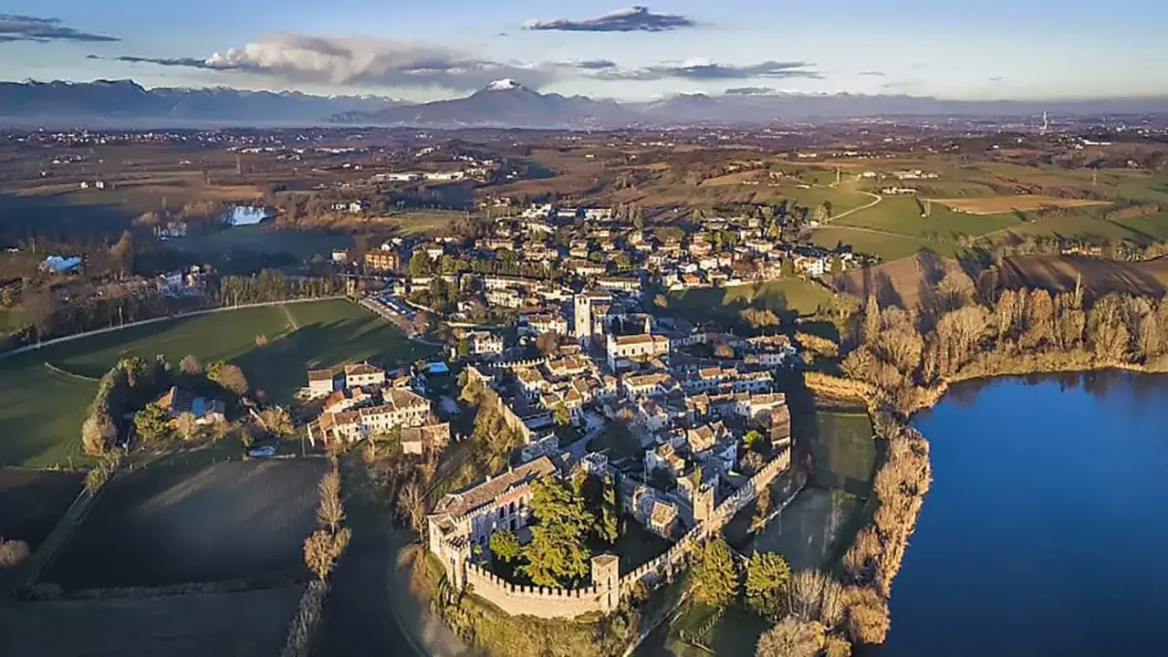 Borgo medievale di Castellaro Lagusello, con mura fortificate, case in pietra e un lago blu, sotto montagne innevate.