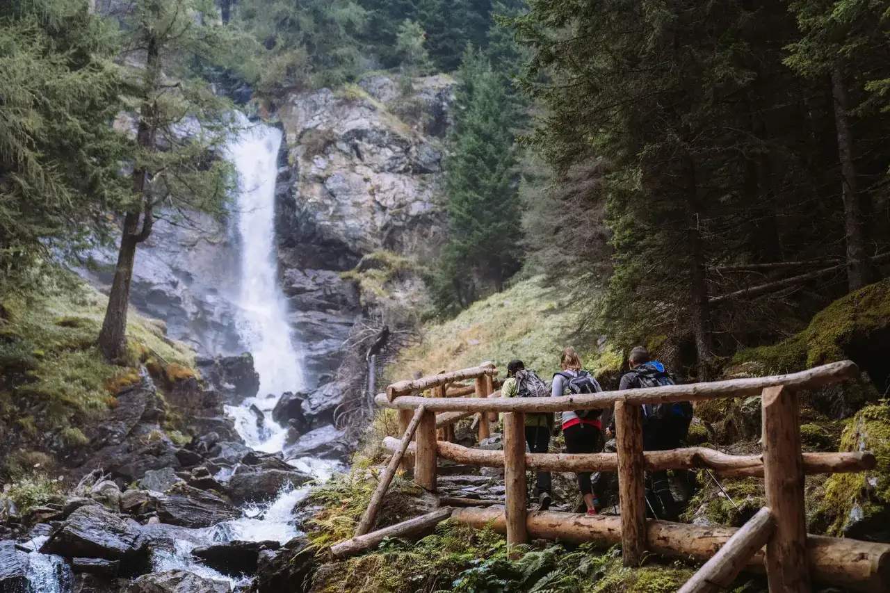 Escursionisti su un sentiero di legno ammirano le maestose cascate di Saent, immerse nel verde di un bosco alpino.