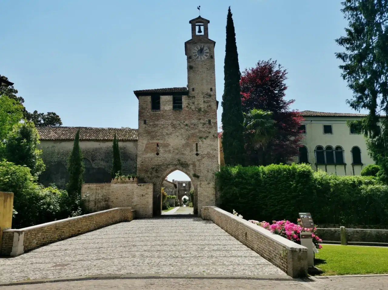 Antica porta con torre dell'orologio, uno dei borghi friulani da visitare, con un ponte in pietra e fiori rosa.