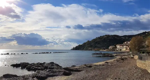 Costa di Pollica con case colorate arroccate sulla collina e mare calmo sotto un cielo nuvoloso.